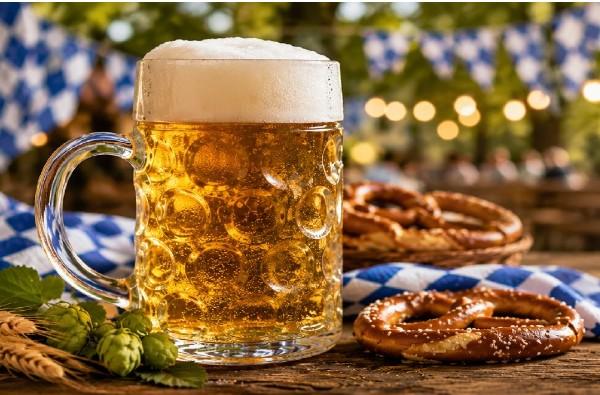 A frothy beer mug beside salted pretzels on a wooden table with Bavarian-themed decorations and blurred festive lights in the background.